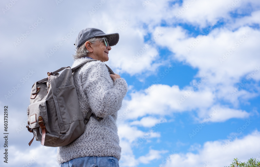 Back view of active senior woman in outdoors walking day holding backpack looking at the blue sky and clouds Back view of active senior woman in outdoors walking day holding backpack looking at the blue sky and clouds