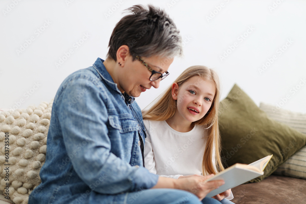 Grandmother and granddaughter spend time together reading a book at home on the sofa.