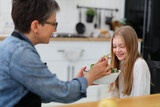 An adult woman feeds salad from a plate to a little girl in the kitchen.