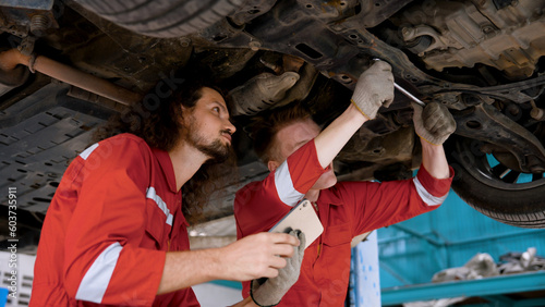 Two mechanic Caucasian man in large garage, Inspecting parts under raised car come in repair problematic part, male holding laptop in hand checking repairs on uncertain issues for correct solution.