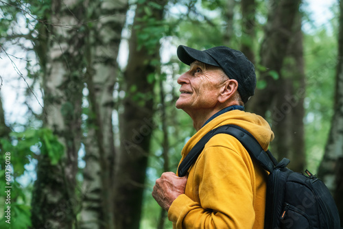 Side view of mature hiker wearing cap and holding backpack standing in summer forest with birch trees. Tourism concept.
