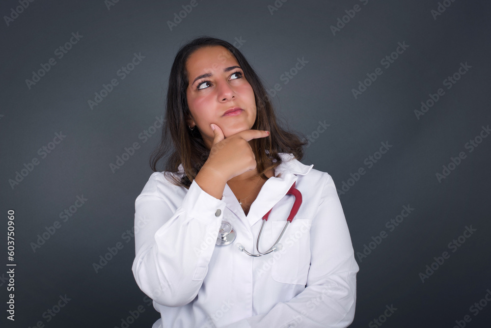 Young beautiful doctor woman wearing medical uniform over gray studio background holding hand under chin having doubtful look. Young beautiful doctor woman wearing medical uniform over gray studio background holding hand under chin having doubtful look.