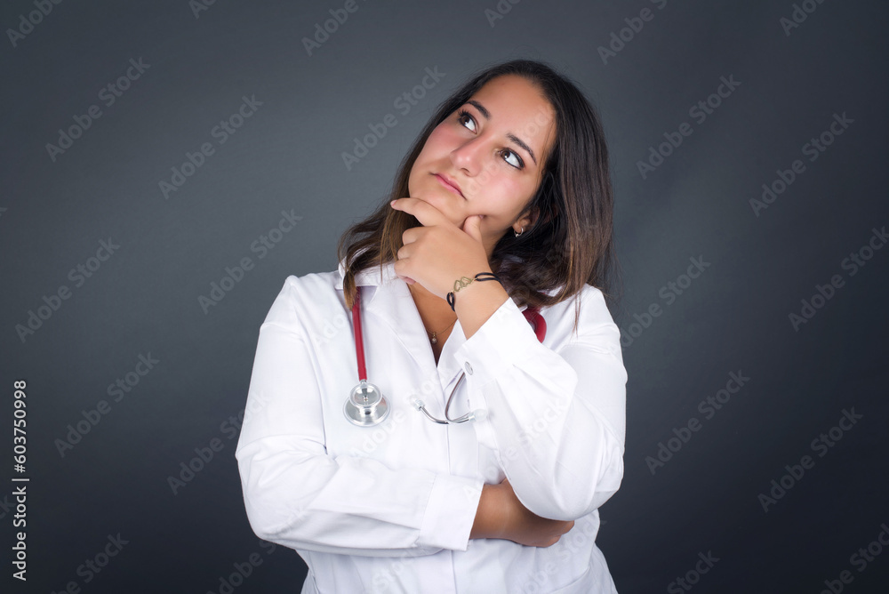 Young beautiful doctor woman wearing medical uniform over gray thinking or wondering about something with interest, dressed casually, poses against gray studio. Taking decisions concept. Young beautiful doctor woman wearing medical uniform over gray thinking or wondering about something with interest, dressed casually, poses against gray studio. Taking decisions concept.