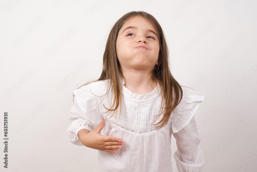Cute charming little girl wearing white shirt over white background puffing cheeks with air. Grimace face, playful kid. 