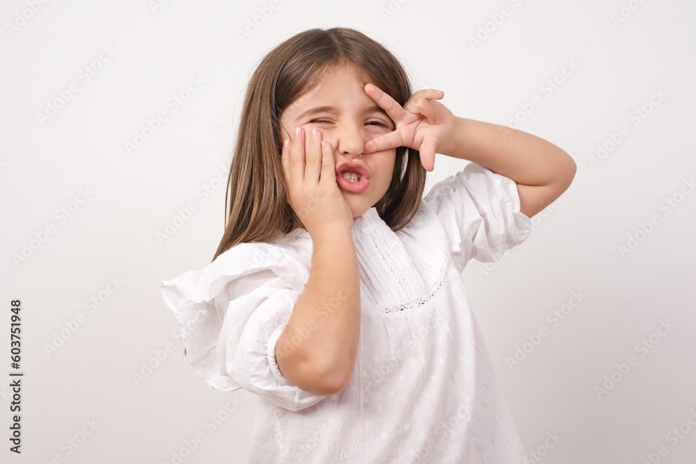 Cute charming little girl wearing white shirt over white background making victory sign near eyes. Cute charming little girl wearing white shirt over white background making victory sign near eyes.