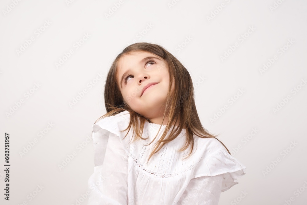 Cute charming little girl wearing white shirt over white background looking up having a dreamy and lovely look. Imagination concept. Cute charming little girl wearing white shirt over white background looking up having a dreamy and lovely look. Imagination concept.