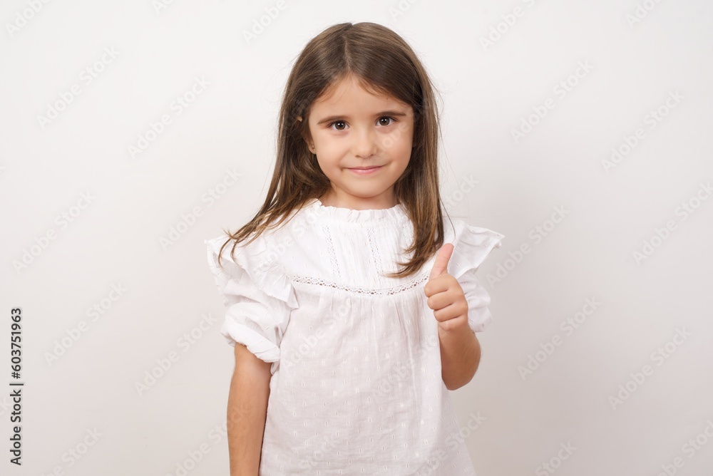 Cute charming little girl wearing white shirt over white background making ok sign with thumb up. Cute charming little girl wearing white shirt over white background making ok sign with thumb up.