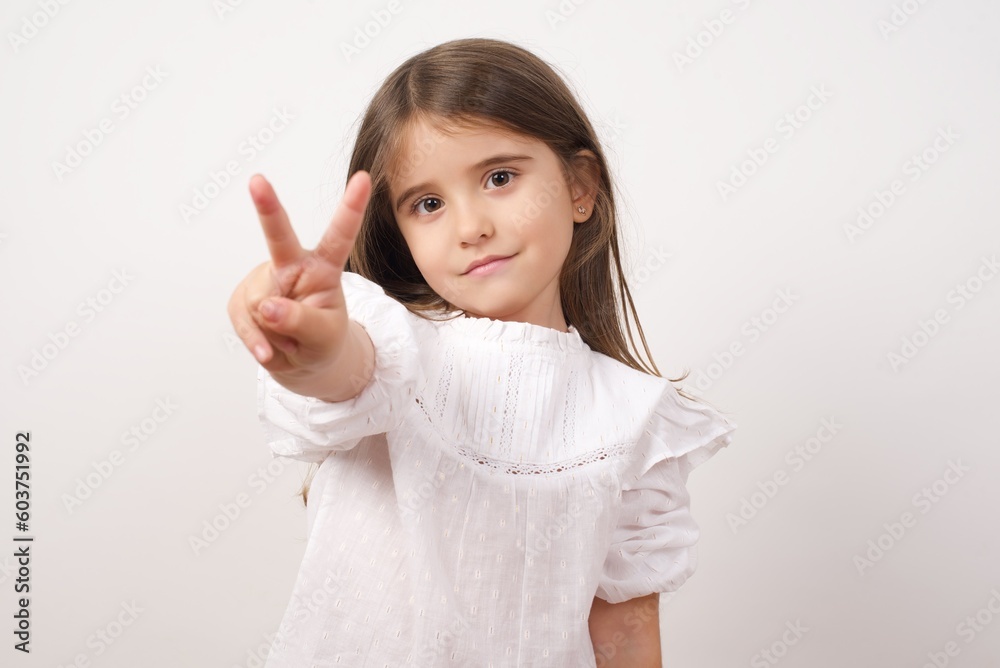 Cute charming little girl wearing white shirt over white background rising two fingers up making Victory sign or number two. Cute charming little girl wearing white shirt over white background rising two fingers up making Victory sign or number two.