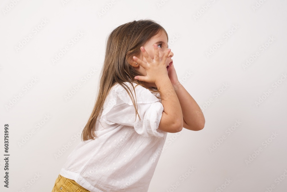 Cute charming little angry girl wearing white shirt over white background looking and screaming aside holding hands near face. Cute charming little angry girl wearing white shirt over white background looking and screaming aside holding hands near face.