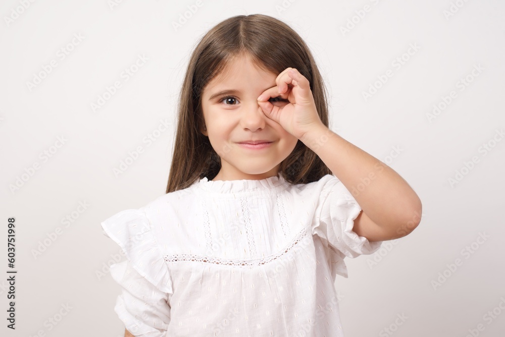 Cute charming little girl wearing white shirt over white background making ok sign with hand on eye. Cute charming little girl wearing white shirt over white background making ok sign with hand on eye.