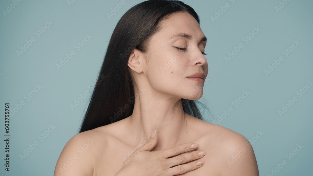 Obraz premium A young woman enjoys her perfect skin by holding her palm on the bare skin under her neck. Close up portrait of woman isolated on blue background. Natural beauty, cosmetology, spa.