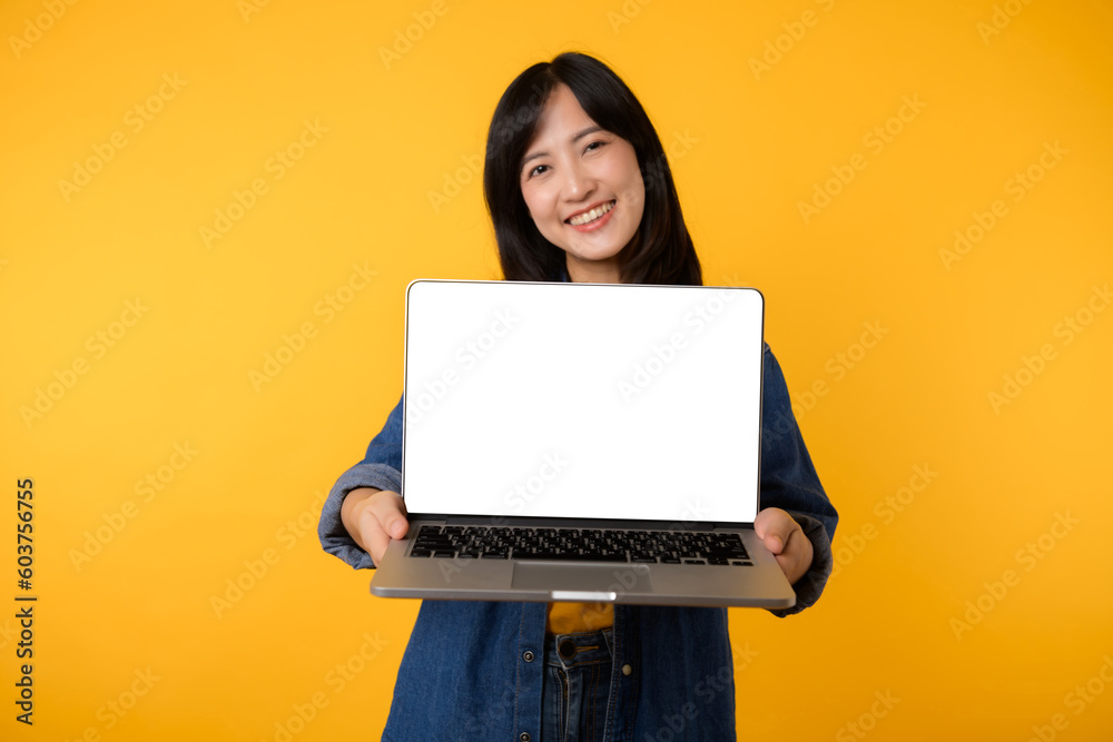 Naklejka premium portrait young happy woman wearing yellow t-shirt and denim shirt holding laptop and point finger to screen isolated on yellow studio background. business technology application communication concept.