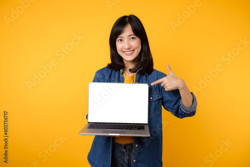 portrait young happy woman wearing yellow t-shirt and denim shirt holding laptop and point finger to screen isolated on yellow studio background. business technology application communication concept.