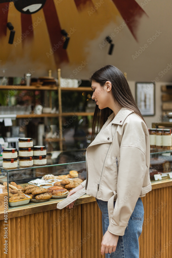 young woman with brunette long hair in beige leather jacket and denim jeans standing near cake display with delicious pastry and jars of jam in modern bakery shop in Istanbul
