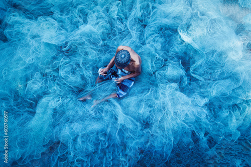 Old fisherman hands sewing blue fishing nets sitting on the ground and surrounded big net photo