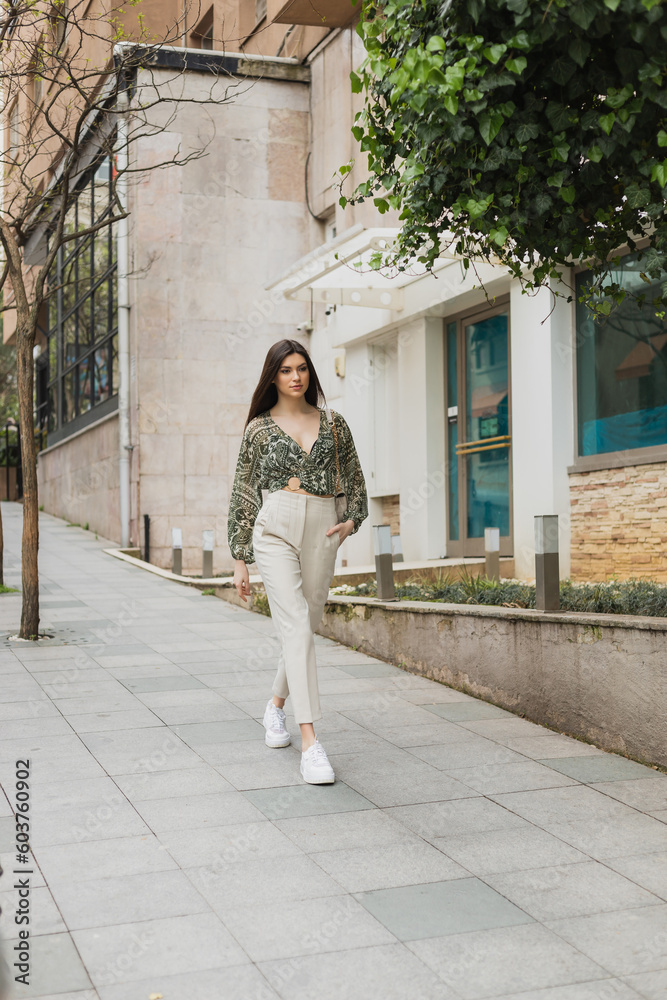 young woman with long hair in trendy outfit with beige pants, cropped blouse and handbag with chain strap walking with hand in pocket near modern building and green tree on street in Istanbul
