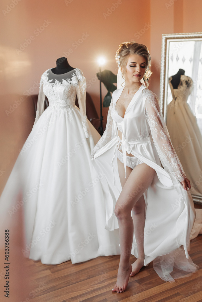 A blonde bride in a long petticoat is standing in her room, posing near her wedding dress. Beautiful hairstyle, open legs. Voluminous dress, open bust