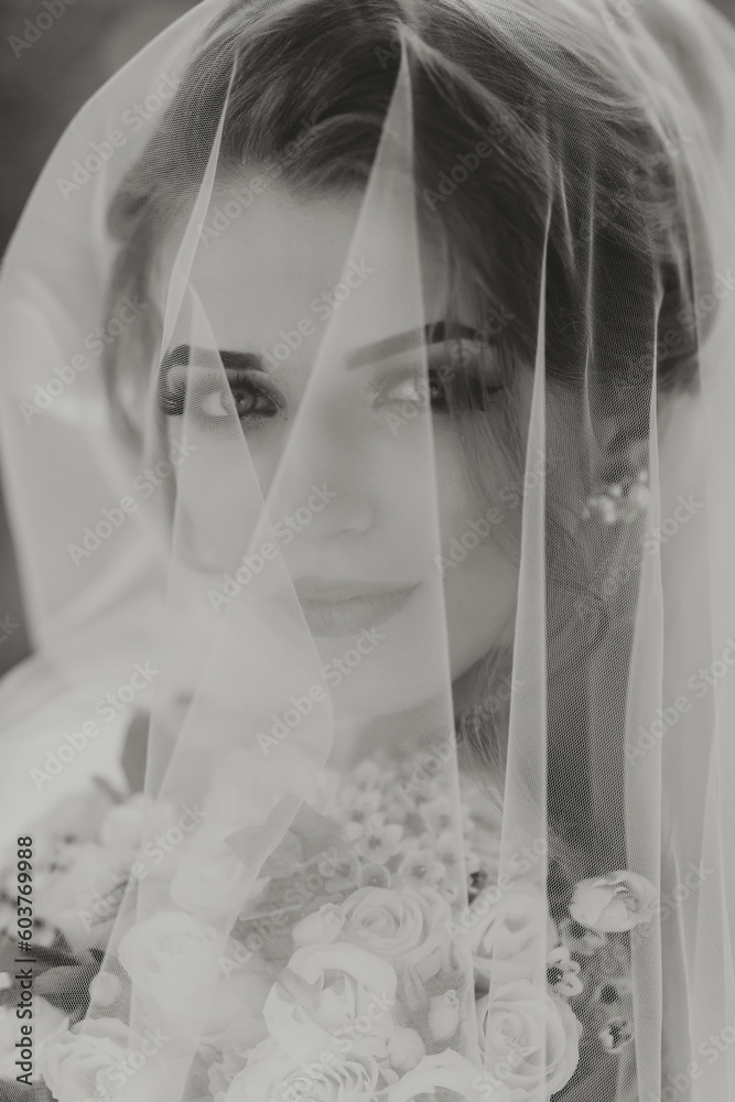 Curly blonde bride in a white dress, covered with a veil, poses for the camera with a bouquet of roses. Portrait of the bride. Beautiful makeup and hair. Wedding in nature. black and white photo Curly blonde bride in a white dress, covered with a veil, poses for the camera with a bouquet of roses. Portrait of the bride. Beautiful makeup and hair. Wedding in nature. black and white photo