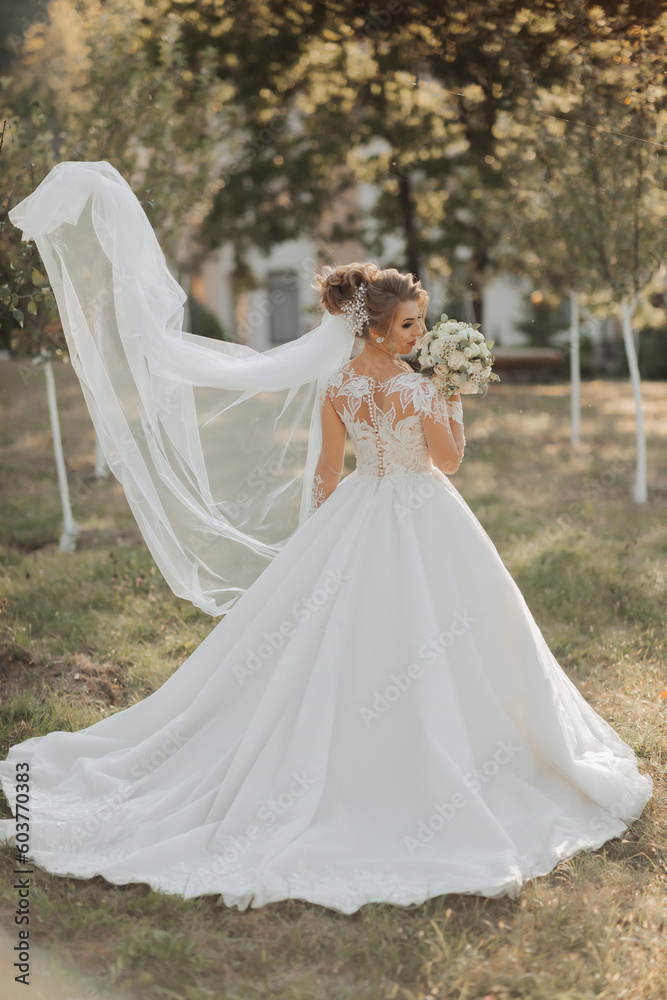 Wedding portrait. A blonde bride in a white dress with a train and lace sleeves holds a bouquet and looks over her shoulder at the camera. A long veil falls in the air. Photo session in nature. Wedding portrait. A blonde bride in a white dress with a train and lace sleeves holds a bouquet and looks over her shoulder at the camera. A long veil falls in the air. Photo session in nature.