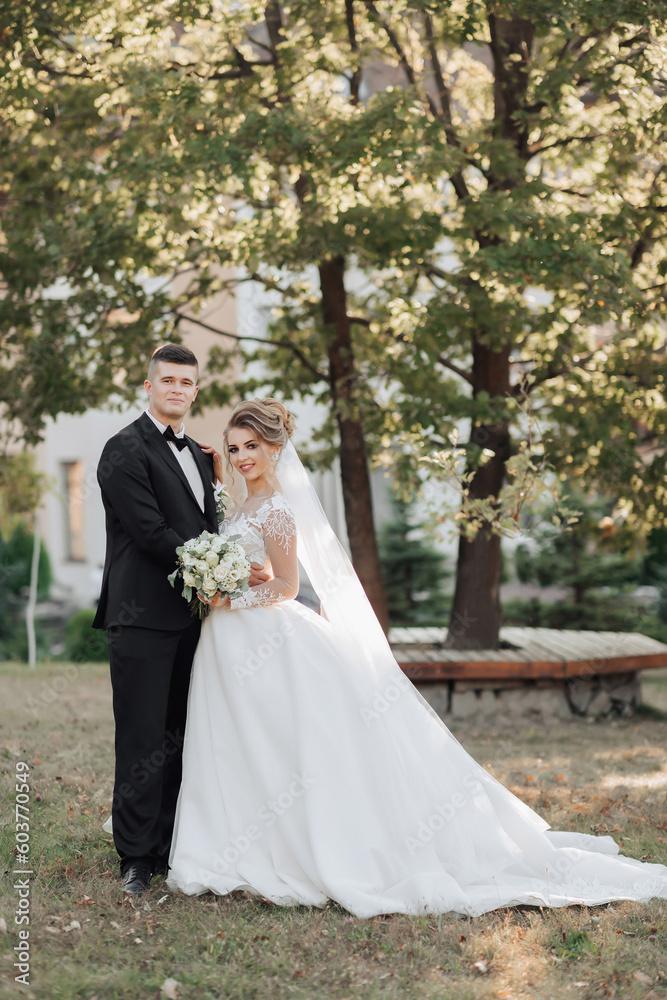 Wedding portrait. The groom in a black suit and the blonde bride are standing, hugging, posing holding a bouquet under a tree. Photo session in nature. Beautiful hair and makeup