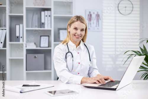 A young female doctor sits in the office in a white coat and with a stethoscope and works on a laptop. Smiling looking at the camera.