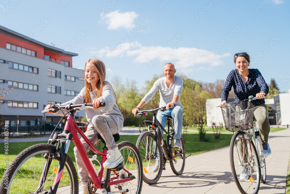 Obraz premium Smiling long-haired little girl with father and mother during summer city outdoor bicycle riding. They enjoy togetherness, happy parenthood and childhood or active sport life concept image