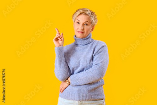 Thoughtful mature woman pointing at something on yellow background