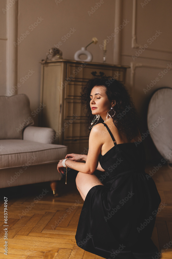 Details of fashion portrait of stylish girl in black dress, sexy pose, beads earrings and necklace. Curly model wearing stylish trendy black long dress sitting on the floor. Fashion outfit. Rear view. Details of fashion portrait of stylish girl in black dress, sexy pose, beads earrings and necklace. Curly model wearing stylish trendy black long dress sitting on the floor. Fashion outfit. Rear view.
