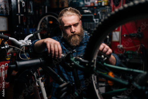 Closeup of handsome bearded technician male repairing and fixing mountain bicycle standing on bike rack working in bike repair shop with dark interior. Concept of maintenance of bicycle transport.