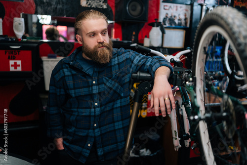 Portrait of thoughtful handsome cycling mechanic male standing by bicycle in repair bike workshop with dark interior, looking away. Concept of professional repair and maintenance of bicycle transport.