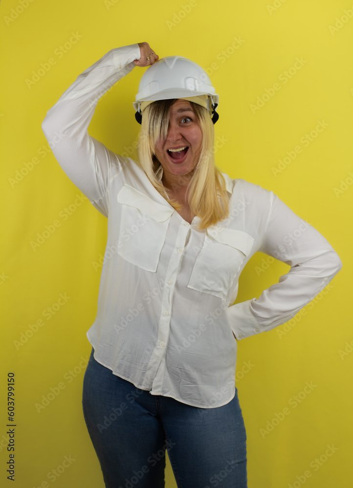 Funny beautiful woman is banging her fist on the construction helmet on her head. A builder woman on a yellow background in a hard hat knocks herself on the head and laughs. Funny beautiful woman is banging her fist on the construction helmet on her head. A builder woman on a yellow background in a hard hat knocks herself on the head and laughs.