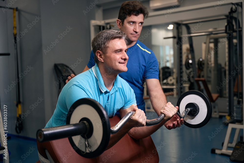Papier Peint photo Lavable Pleased adult male performing bicep strength  exercise with personal trainer 