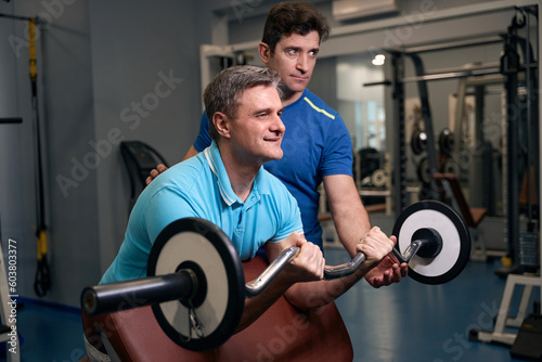 Fototapete Pleased adult male performing bicep strength exercise with personal trainer