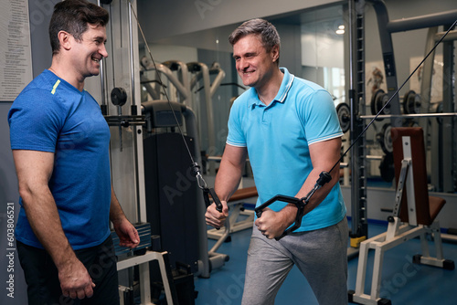 Smiling male working out on cable machine supervised by coach