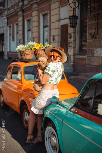 Young woman sitting on the hood of a retro car on a summer day playing with her puppy © Julia