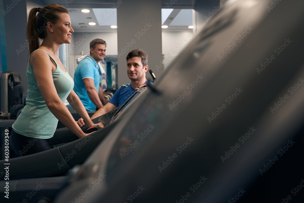 Smiling gym-goer exercising on cardio machine supervised by fitness coach