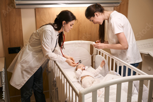 Serious pediatrician auscultating newborn baby in hospital ward