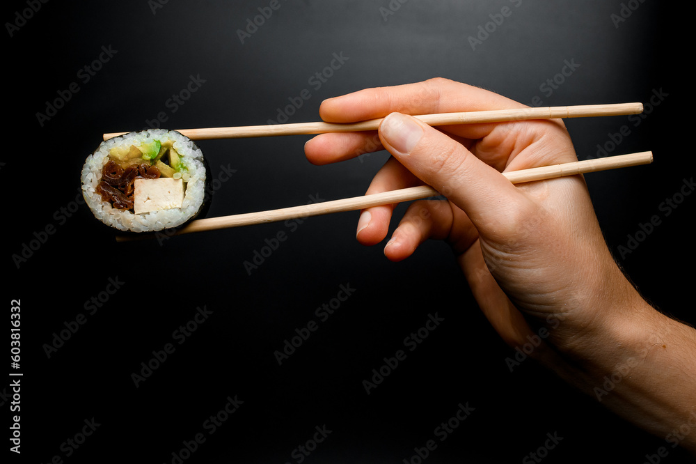 Man's hand holds tasty sushi roll with smoked eel, avocado, cucumber and cheese using chopsticks Man's hand holds tasty sushi roll with smoked eel, avocado, cucumber and cheese using chopsticks