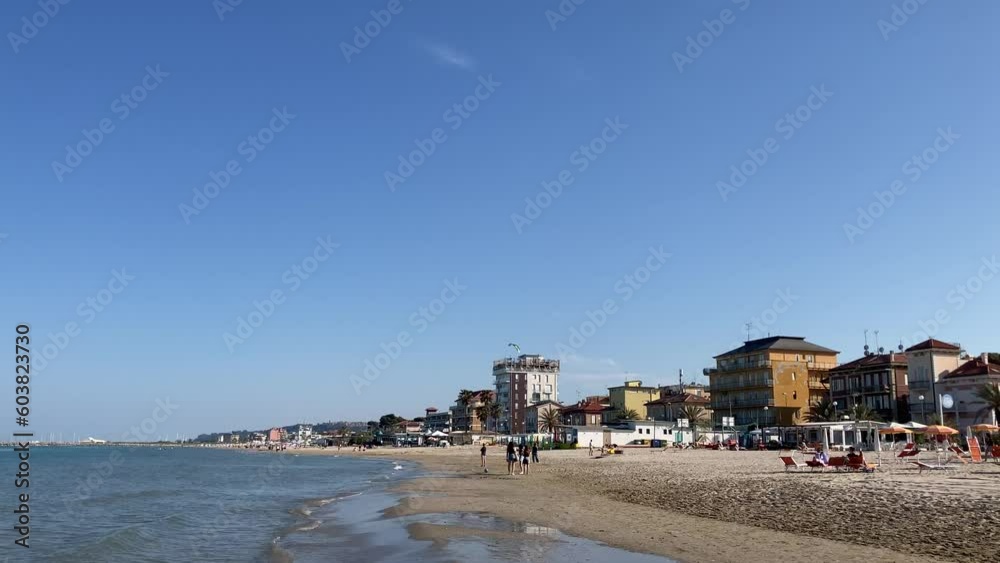 Intriguing panorama in Porto San Giorgio on the Adriatic Sea with a paraglider in the sky above the beach circling a black and white building with different onlookers, parasols and sun loungers Intriguing panorama in Porto San Giorgio on the Adriatic Sea with a paraglider in the sky above the beach circling a black and white building with different onlookers, parasols and sun loungers