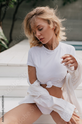 Elegant pretty stylish long woman wearing white top and shirt is posing near the pool with closed eyes and tender smile 