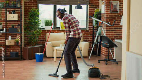 African american man dancing and vacuuming home floors, singing and listening to music on headset. Male person having fun spring cleaning, using vacuum to clean dust and dirt.