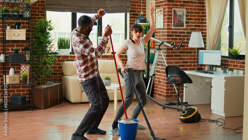 Happy man and woman doing house chores and dancing, listening to music for spring cleaning work. Cheerful couple sweeping floors and using vacuum, singing together in living room.