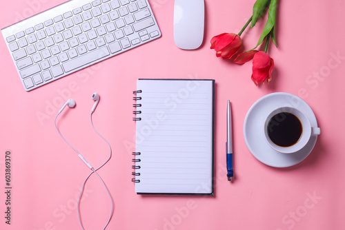 Flat lay home office desk. Business woman workspace with blank notebook for text, keyboard, smartphone, alarm clock, red roses and coffee cup on pink background. Top view feminine background.