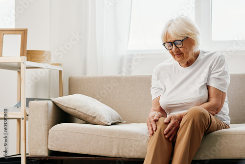 Elderly woman severe pain in her leg sitting on the couch, health problems in old age, poor quality of life. Grandmother with gray hair holds on to her sore knee, problems with joints and ligaments.