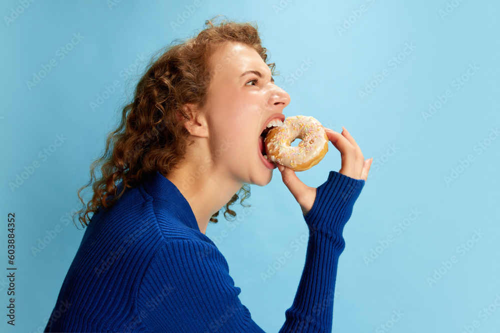 Portrait of young curly girl in blue top emotional eating donut against blue studio background. Sweet tooth. Concept of human emotions, youth culture, fashion, lifestyle, food Portrait of young curly girl in blue top emotional eating donut against blue studio background. Sweet tooth. Concept of human emotions, youth culture, fashion, lifestyle, food