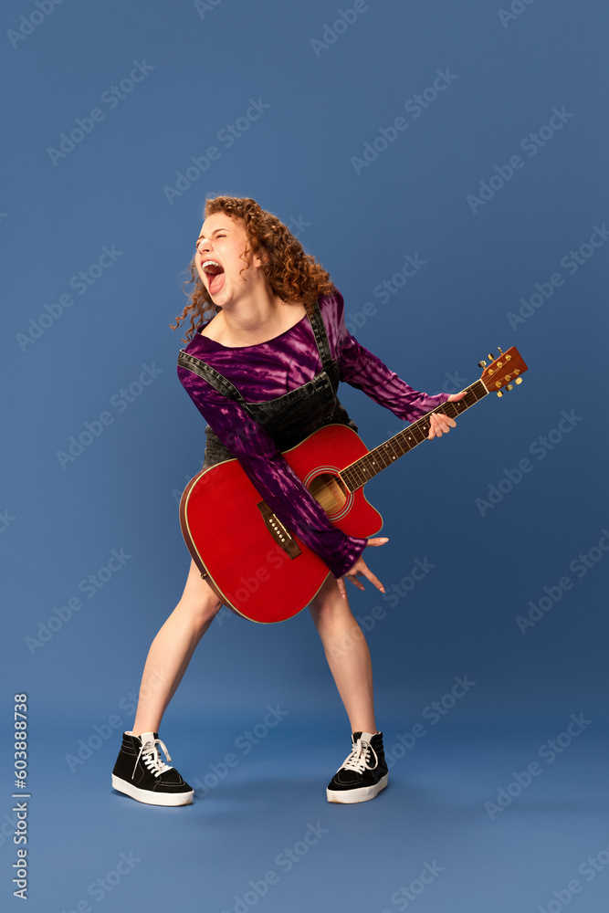 Shouting, singing. Burst of energy. Portrait of young emotional girl playing guitar, having fun against blue studio background. Concept of human emotions, youth culture, fashion, creative lifestyle Shouting, singing. Burst of energy. Portrait of young emotional girl playing guitar, having fun against blue studio background. Concept of human emotions, youth culture, fashion, creative lifestyle