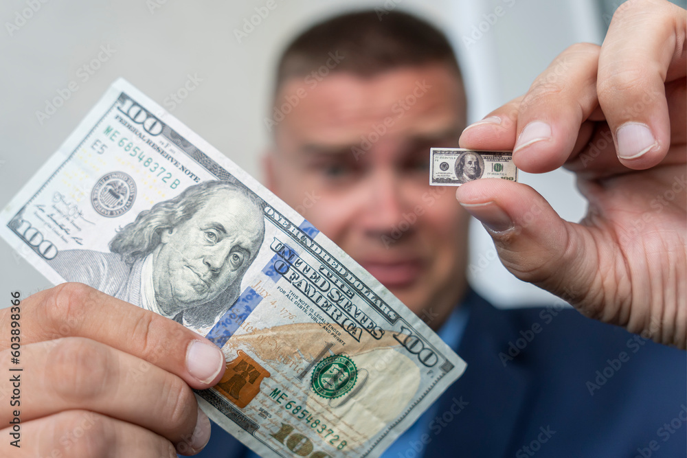 A young businessman guy holds a small bill in his hand as a symbol of declining income. Small salary concept. A sad face . dollar inflation A young businessman guy holds a small bill in his hand as a symbol of declining income. Small salary concept. A sad face . dollar inflation