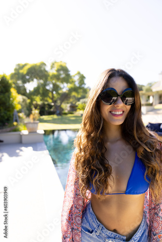 Portrait of smiling fit caucasian woman in sunglasses standing by pool in sunny garden, copy space