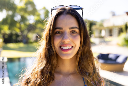 Portrait of smiling caucasian fit woman with sunglasses on head standing by pool in sunny garden