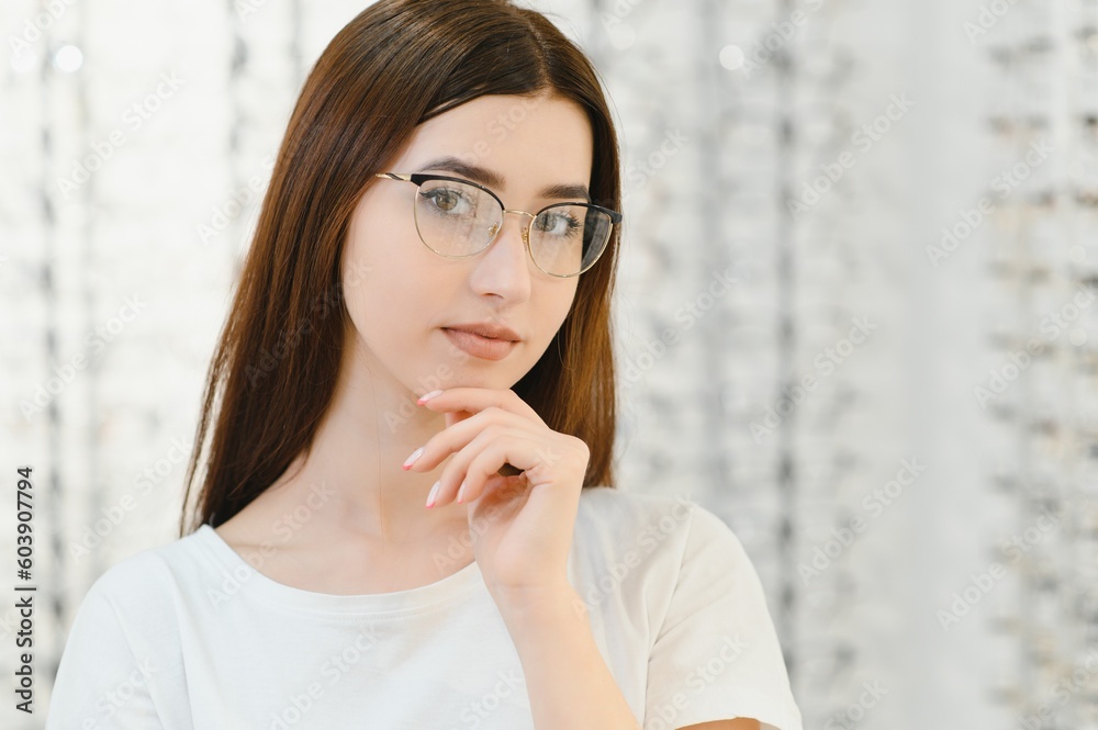 Young girl trying on eyeglasses in front of mirror. Helping to improve vision, saving eye health. Standing near transparent stand with optical lenses. Young girl trying on eyeglasses in front of mirror. Helping to improve vision, saving eye health. Standing near transparent stand with optical lenses.